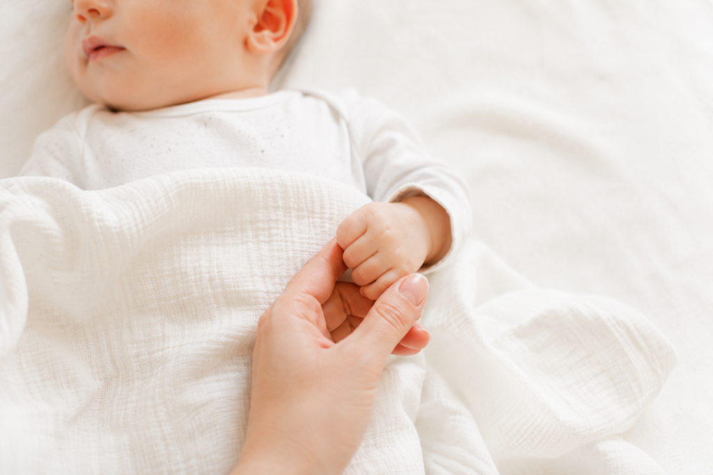 Bébé allongé sur une couverture blanche tenant le doigt d'un adulte dans une ambiance lumineuse, douce et épurée.