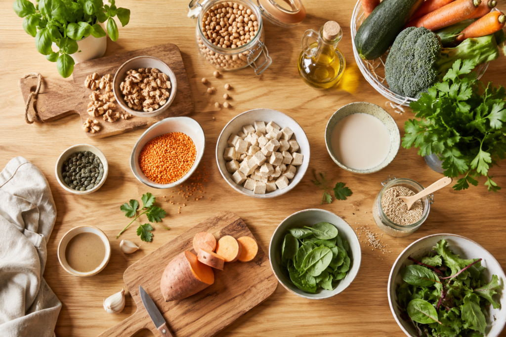 Vue de dessus d'un plan de travail en bois clair avec des légumes frais, des légumineuses, des graines, une huile végétale et des alternatives végétales dans une cuisine chaleureuse.